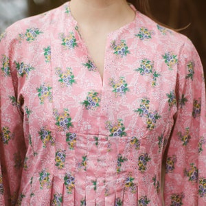 May include: A pink floral print dress with a mandarin collar and long sleeves. The dress features a V-neckline and pleats at the waist. The floral pattern includes blue, yellow, and green flowers on a white lace-like background.