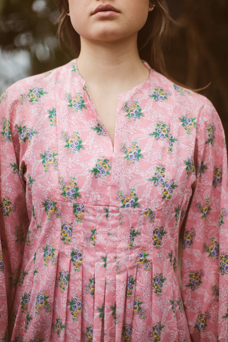 May include: A pink floral print dress with a mandarin collar and long sleeves. The dress features a V-neckline and pleats at the waist. The floral pattern includes blue, yellow, and green flowers on a white lace-like background.