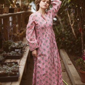May include: A long, pink floral dress with long sleeves and a V-neck. The dress features a pattern of small blue and yellow flowers. The person is standing in a greenhouse with wooden shelves and potted plants.