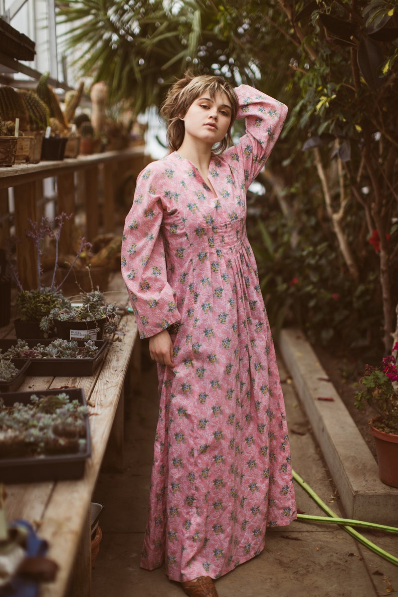 May include: A long, pink floral dress with long sleeves and a V-neck. The dress features a pattern of small blue and yellow flowers. The person is standing in a greenhouse with wooden shelves and potted plants.