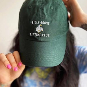 May include: A dark green baseball cap with the text "SILLY GOOSE LIFTING CLUB" embroidered on the front. The cap features a white cartoon goose lifting weights. The cap is made of a soft material.