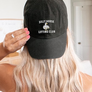 May include: Black baseball cap with the text "SILLY GOOSE LIFTING CLUB" and a cartoon goose lifting weights. The cap is made of a soft fabric and has a curved brim. The cap is a great accessory for anyone who enjoys lifting weights.