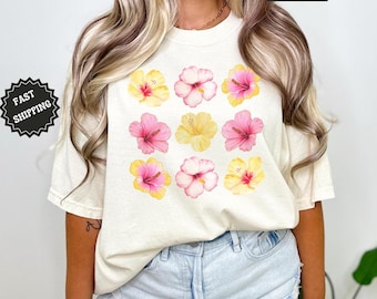 May include: A woman is wearing a cream colored t-shirt with a floral print of pink and yellow hibiscus flowers. The shirt has the text "COMFORT COLORS" printed on the top. The woman is wearing light blue denim shorts.