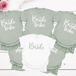 May include: Five t-shirts in various shades of green and white with the text "Bride Tribe" printed on them. The shirts are arranged in a semi-circle with the white shirt in the center.