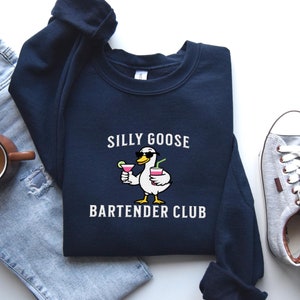 May include: A navy blue sweatshirt featuring a white graphic of a cartoon goose wearing sunglasses and holding two cocktails. The text "Silly Goose Bartender Club" is printed below the goose.