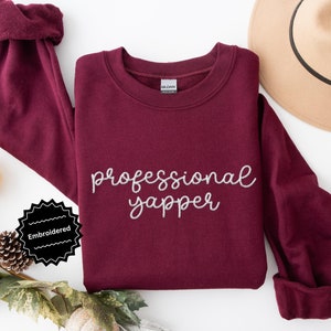 May include: Burgundy sweatshirt with the text "professional yapper" embroidered in silver thread. The sweatshirt is laid flat on a white surface with a small pine cone and a pumpkin in the background.