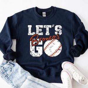 May include: Navy blue sweatshirt with a white distressed graphic that says "Let's Go Broncos" with a baseball graphic. The sweatshirt is laying on a pair of white sneakers and light blue jeans.