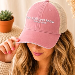 May include: A dusty rose baseball cap with a mesh back. The cap features the embroidered text "be still and know" and "Psalm 46:10" in white. The brim has a floral pattern. A succulent plant in a terracotta pot is in the background.