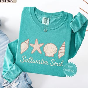 May include: Seafoam green long-sleeve shirt with a seashell, starfish, and shell graphic. The text "Saltwater Soul" is printed below the graphic. The shirt is made of a soft material and is a comfortable fit.