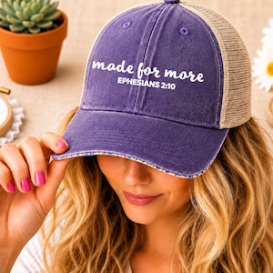 May include: A purple baseball cap with a mesh back and the words "made for more" and "Ephesians 2:10" printed on the front. The cap has a curved brim and a button on top. The cap is a trucker style hat.