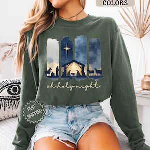 May include: Green long-sleeve sweatshirt with a Christmas nativity scene design. The design features a star, stable, and silhouettes of figures and animals. The words "oh holy night" are written in a script font. The sweatshirt has the words "Comfort Colors" and "Hemp" printed on it.