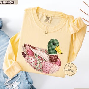 May include: A light yellow long-sleeve shirt with a patchwork duck design. The duck features a green checkered head, a yellow beak, and wings with floral and heart patterns. The shirt has the words "COMFORT COLORS" and "BUTTER" printed on it.