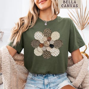 May include: An olive green t-shirt featuring a floral design made from various fabric patterns, including leopard print and gingham. The text "BELLA CANVAS" is in the upper right corner, and "MILITARY" is at the bottom. The t-shirt is worn with denim shorts.
