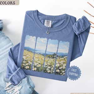 May include: A blue long-sleeve shirt with a landscape design featuring a field of white flowers and a blue sky. The shirt has the text "Comfort Colors" on the upper left and a silver necklace. The shirt is folded next to a pair of blue jeans.