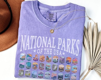 Könnte beinhalten: Ein lavendelfarbenes T-Shirt mit einer weißen Grafik, auf der "National Parks of the USA" steht und eine Sammlung bunter Nationalpark-Logos abgebildet ist.