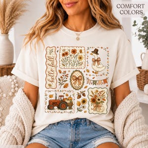 May include: An ivory t-shirt featuring a graphic print with the text 'hello fall' and various autumn-themed illustrations including a red tractor, a goose wearing a hat, floral arrangements, pumpkins, and a plaid bow within decorative frames.