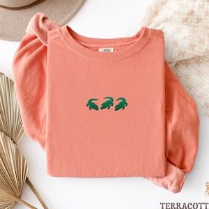 May include: A terracotta-colored long-sleeve shirt with three green alligator designs embroidered across the chest. The shirt is folded, with the sleeves visible. The word "TERRACOTTA" is at the bottom of the image.