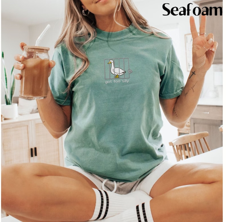 May include: Seafoam green t-shirt with a cartoon duck in a cage design. The duck is white with a yellow beak and feet. The text "got too silly" is below the duck. The shirt is a relaxed fit.