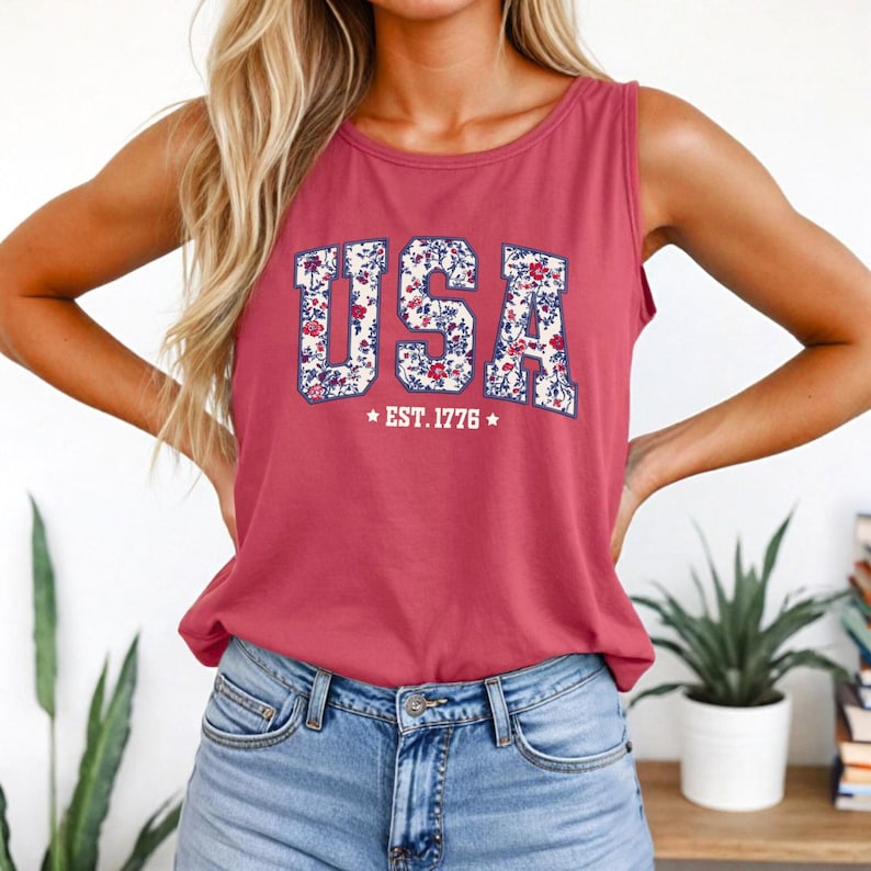 K&ouml;nnte beinhalten: Ein weinrotes Tanktop mit dem Wort "USA" in einem floralen Design. Der Text "EST. 1776" steht unter dem Wort "USA". Das Tanktop wird mit blauen Jeans getragen.