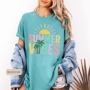 K&ouml;nnte beinhalten: Ein t&uuml;rkisfarbenes T-Shirt mit den Worten "SUMMER VIBES" in bunten Buchstaben, einer gelben Sonne und zwei Palmen. Das Shirt ist Teil einer sommerlichen Bekleidungskollektion.