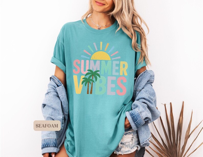 K&ouml;nnte beinhalten: Ein t&uuml;rkisfarbenes T-Shirt mit den Worten "SUMMER VIBES" in bunten Buchstaben, einer gelben Sonne und zwei Palmen. Das Shirt ist Teil einer sommerlichen Bekleidungskollektion.