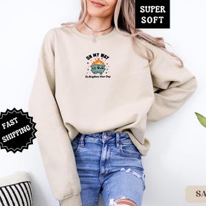 Pu&ograve; includere: Una felpa color sabbia con una grafica di un cestino in fiamme con la scritta "Oh My Way To Brighten Your Day". La felpa &egrave; etichettata "Super Soft" e ha un adesivo "Fast Shipping".
