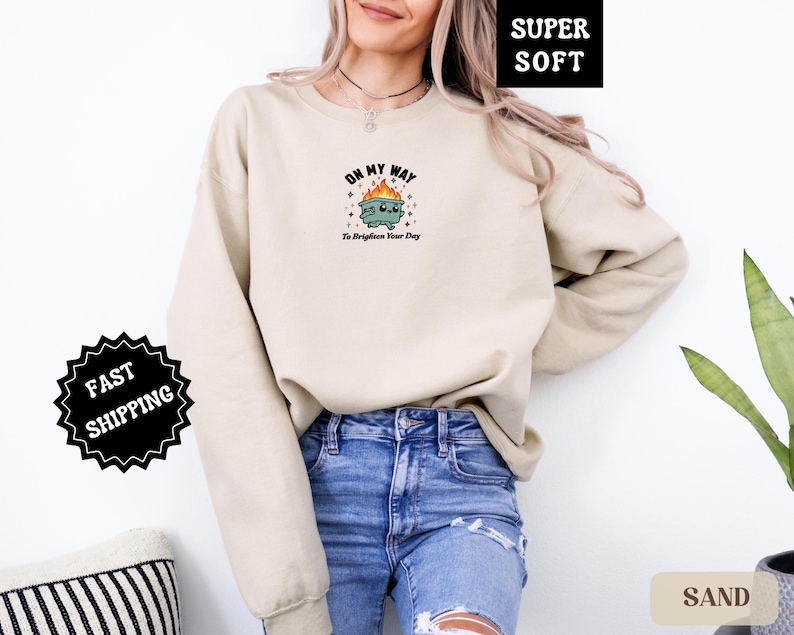 Pu&ograve; includere: Una felpa color sabbia con una grafica di un cestino in fiamme con la scritta "Oh My Way To Brighten Your Day". La felpa &egrave; etichettata "Super Soft" e ha un adesivo "Fast Shipping".