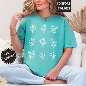 May include: A teal blue t-shirt with a white floral design. The shirt has the text "Comfort Colors" printed on the top left corner.