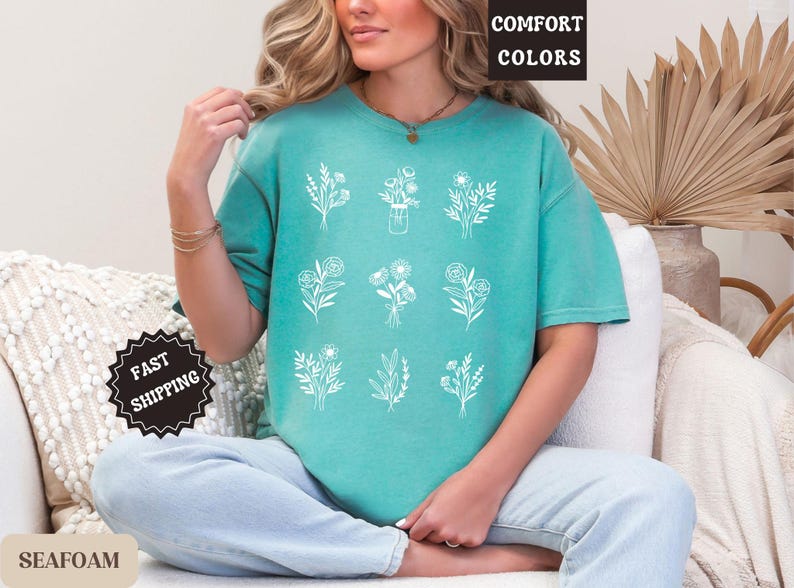 May include: A teal blue t-shirt with a white floral design. The shirt has the text "Comfort Colors" printed on the top left corner.