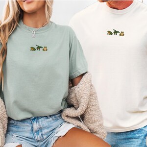 May include: Two t-shirts, one in bay green and the other in ivory, each featuring an embroidered design of three small frogs. The bay green shirt is paired with denim shorts, while the ivory shirt is worn with blue jeans.