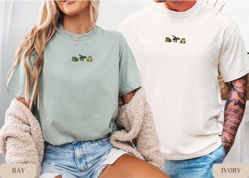 May include: Two t-shirts, one in bay green and the other in ivory, each featuring an embroidered design of three small frogs. The bay green shirt is paired with denim shorts, while the ivory shirt is worn with blue jeans.