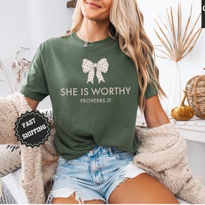 May include: Moss green t-shirt with the text "SHE IS WORTHY PROVERBS 31" and a bow graphic. The shirt is a Comfort Colors brand. A "Fast Shipping" sticker is visible.