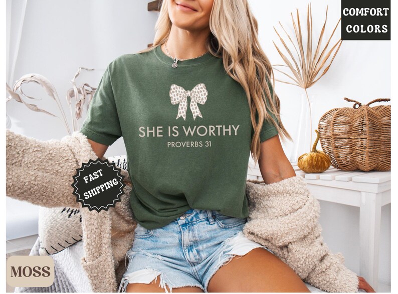 May include: Moss green t-shirt with the text "SHE IS WORTHY PROVERBS 31" and a bow graphic. The shirt is a Comfort Colors brand. A "Fast Shipping" sticker is visible.