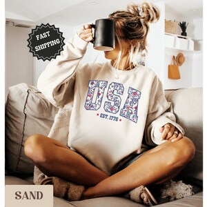 Peut inclure: Un sweat-shirt couleur sable avec le mot "USA" dans un motif floral, avec "EST. 1776" en dessous. Le sweat-shirt est port&eacute; par une personne tenant une tasse noire.