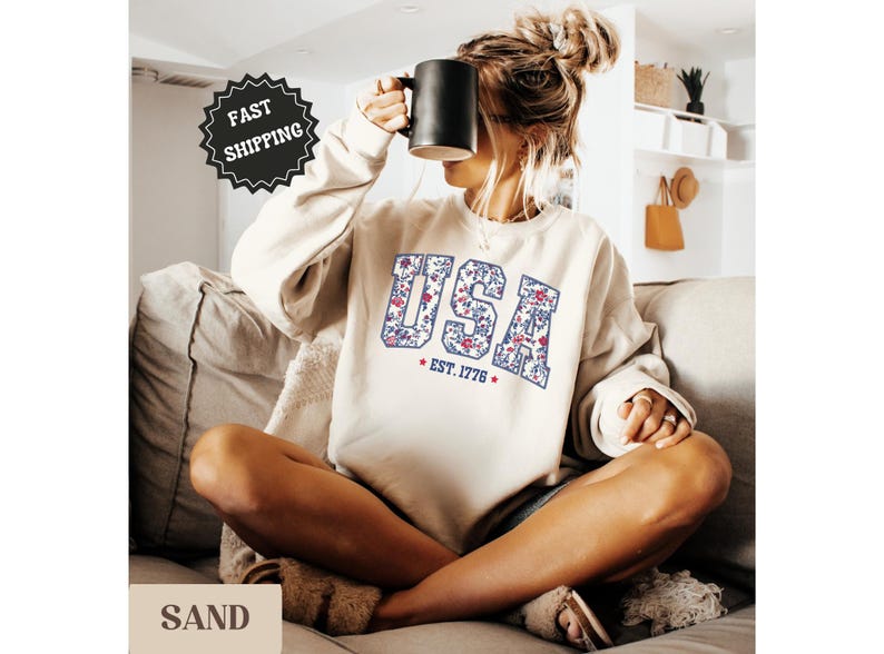 Peut inclure: Un sweat-shirt couleur sable avec le mot "USA" dans un motif floral, avec "EST. 1776" en dessous. Le sweat-shirt est port&eacute; par une personne tenant une tasse noire.