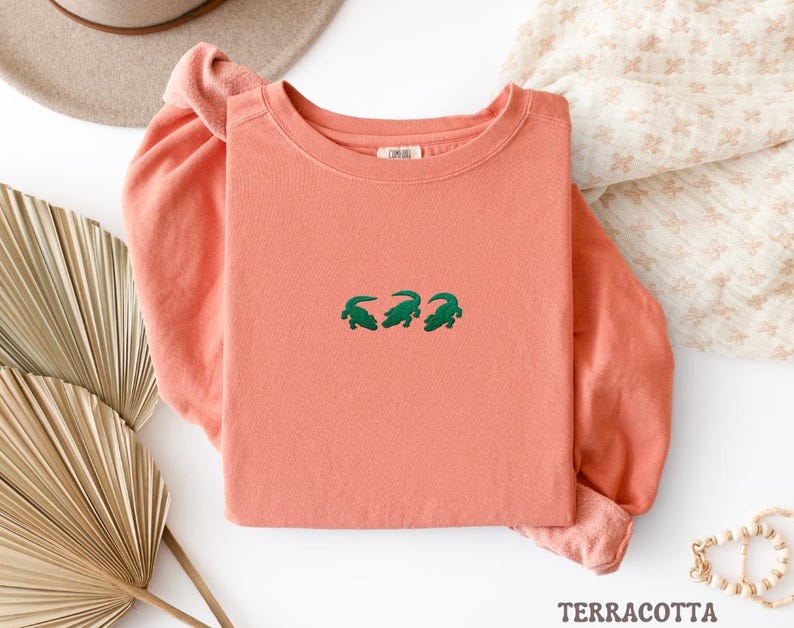May include: A terracotta-colored long-sleeve shirt with three green alligator designs embroidered across the chest. The shirt is folded, with the sleeves visible. The word "TERRACOTTA" is at the bottom of the image.