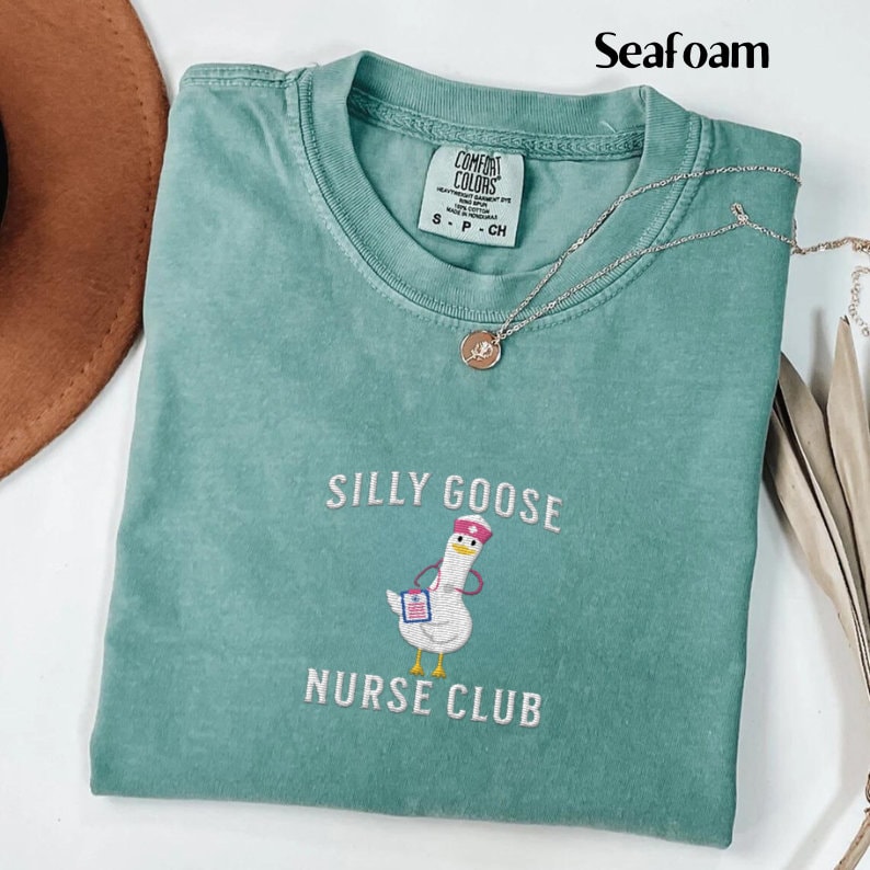 Puede incluir: Camiseta verde espuma de mar con dise&ntilde;o bordado "Silly Goose Nurse Club". El dise&ntilde;o presenta un ganso de dibujos animados con gorro y uniforme de enfermera. Un collar plateado con colgante est&aacute; sobre la camiseta.