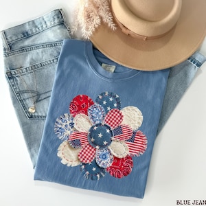 May include: A pale blue t-shirt featuring a flower design made from red, white, and blue fabric patches. The shirt is styled with light wash blue jeans and a beige hat. A silver necklace is also visible.