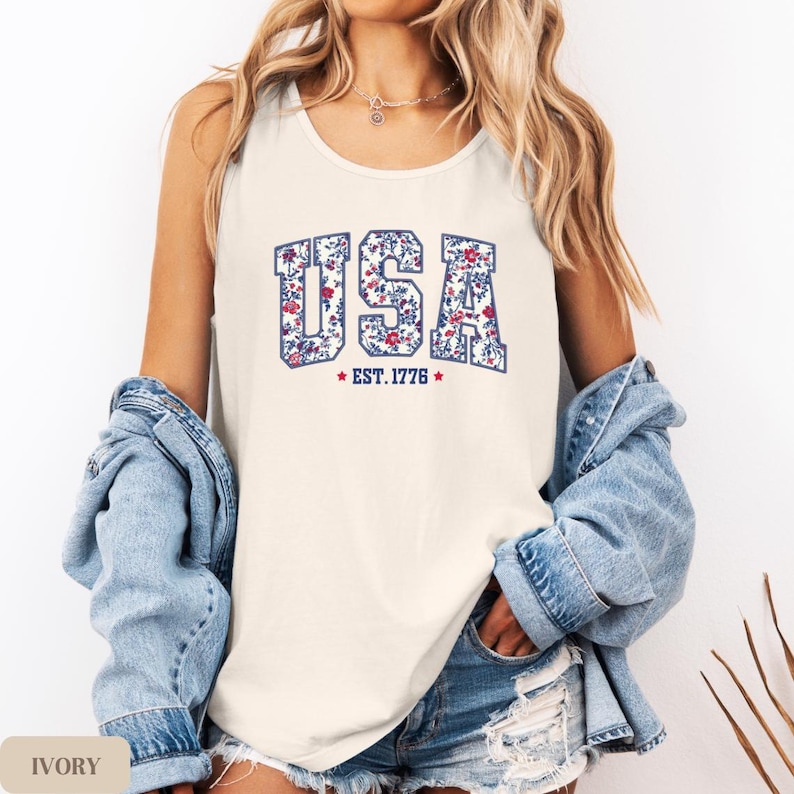 K&ouml;nnte beinhalten: Elfenbeinfarbenes Tanktop mit dem Wort "USA" in floralem Design und "EST. 1776" darunter. Das Tanktop wird mit einer Jeansjacke und Shorts getragen.