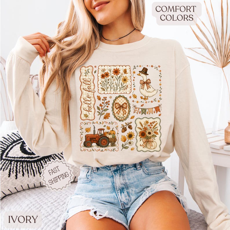 May include: An ivory long-sleeve sweatshirt featuring a graphic print with the text "hello fall". The design includes a tractor, a goose wearing a hat, sunflowers, autumn leaves, a bow, and bunting. The sweatshirt has a crew neckline.