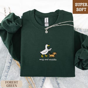 May include: Forest green sweatshirt with the text "wag and waddle" and a graphic of a duck walking a dog. The sweatshirt is labeled "Super Soft". A silver necklace is draped across the sweatshirt.