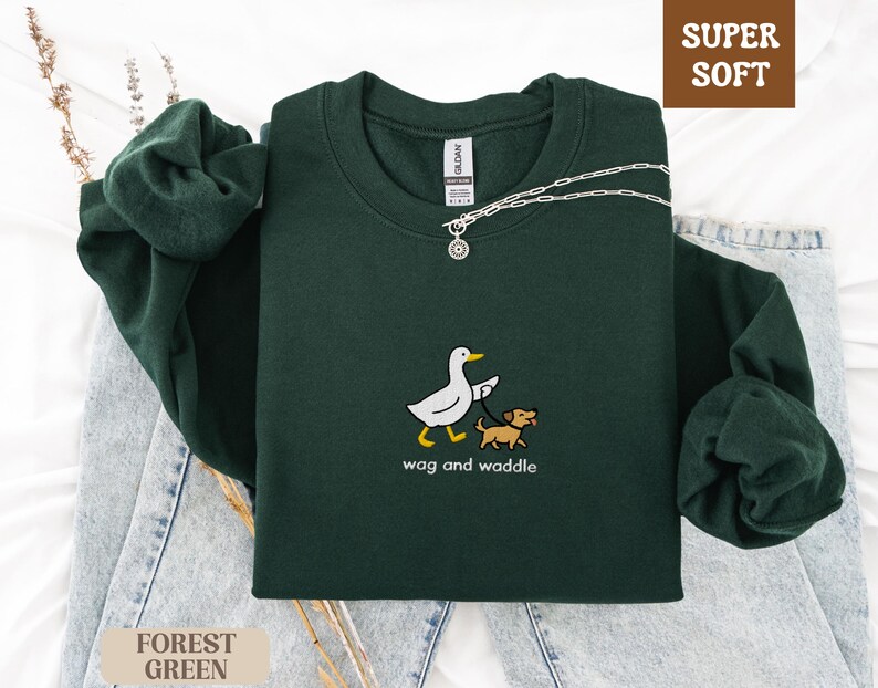 May include: Forest green sweatshirt with the text "wag and waddle" and a graphic of a duck walking a dog. The sweatshirt is labeled "Super Soft". A silver necklace is draped across the sweatshirt.