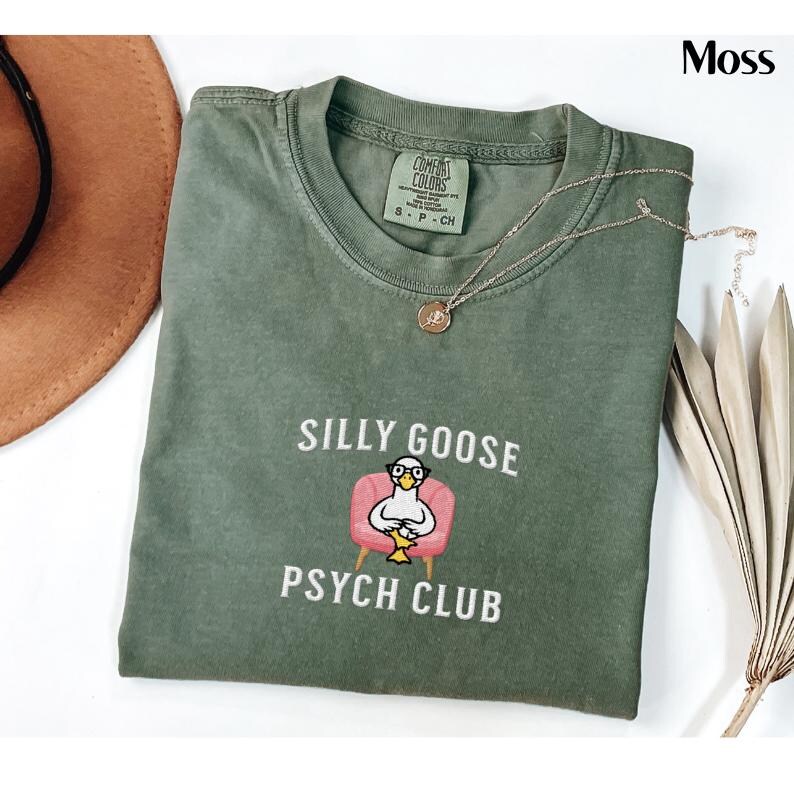 May include: A green t-shirt with the text "Silly Goose Psych Club" and a cartoon goose wearing glasses sitting in a pink armchair.