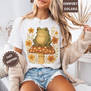 May include: White t-shirt featuring a green frog perched on a mushroom with yellow flowers. The text "COMFORT COLORS" appears in the top right corner of the image.