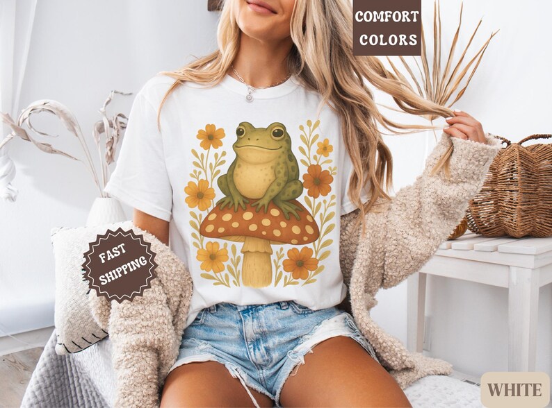 May include: White t-shirt featuring a green frog perched on a mushroom with yellow flowers. The text "COMFORT COLORS" appears in the top right corner of the image.