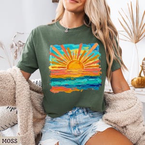 Puede incluir: Camiseta verde musgo con un dise&ntilde;o vibrante de puesta de sol. La obra de arte representa un sol con rayos naranjas y amarillos sobre un oc&eacute;ano colorido. La camiseta es de estilo casual y c&oacute;modo.