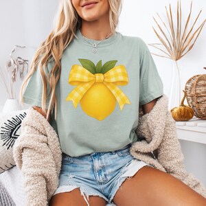 Puede incluir: Una camiseta azul claro con un lim&oacute;n amarillo con un lazo de cuadros amarillos y blancos en la parte superior. El lim&oacute;n tiene hojas verdes.