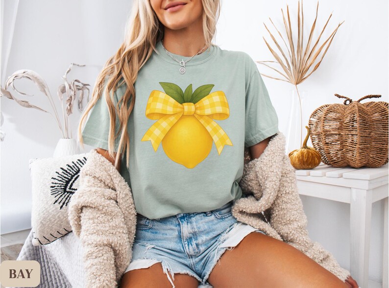 Puede incluir: Una camiseta azul claro con un lim&oacute;n amarillo con un lazo de cuadros amarillos y blancos en la parte superior. El lim&oacute;n tiene hojas verdes.