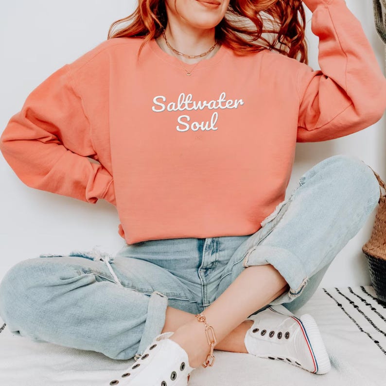 Op de afbeelding: Koraalrood sweatshirt met de tekst "Saltwater Soul" in witte letters. Het sweatshirt wordt gecombineerd met lichtblauwe jeans en witte sneakers. De algehele uitstraling is casual en comfortabel.