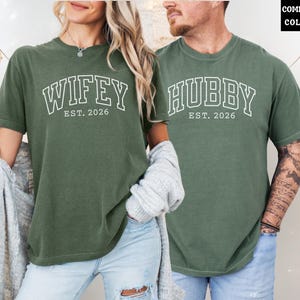 May include: Two moss green t-shirts. One says "Wifey Est. 2026" in white block letters, the other says "Hubby Est. 2026". The shirts are worn by a couple. The background is neutral with decorative lights.